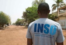 A man stands on a dirt road wearing a gray INSO t-shirt, surrounded by trees and buildings under a clear sky.