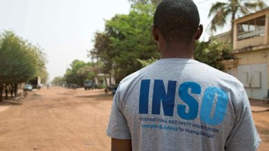 A man stands on a dirt road wearing a gray INSO t-shirt, surrounded by trees and buildings under a clear sky.