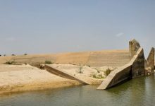 Concrete dam ruins with slanted walls near a calm waterbody, surrounded by sand and sparse vegetation under a clear sky.