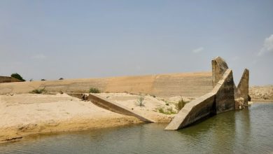 Concrete dam ruins with slanted walls near a calm waterbody, surrounded by sand and sparse vegetation under a clear sky.