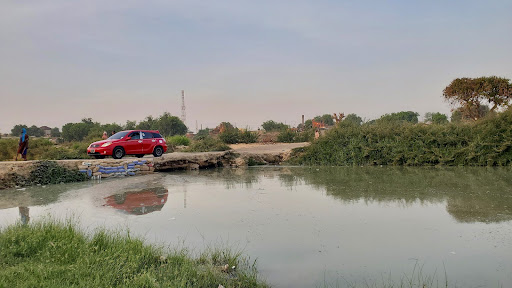 A red car is parked on a narrow bridge over a calm river with greenery and a person nearby under a clear sky.