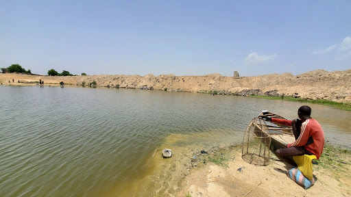 Person sitting near a pond with fishing gear, looking at the water under a clear sky.