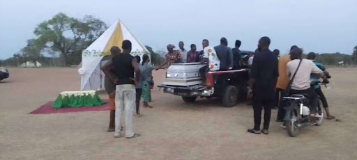 A group of people gather around a tent and a pickup truck carrying a coffin on a field, with a motorcycle nearby.