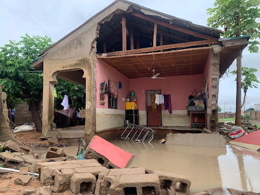 Flooded house with damaged walls and furniture inside. Debris and water cover the floor. Tree visible in the background.