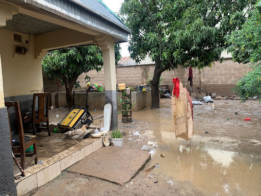 Flooded yard with scattered items and muddy water. A duck and trees are visible near a house with a partially covered porch.