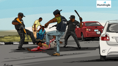 Police officers in tactical gear confront and restrain a person on the road while others observe, with vehicles in the background.