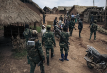 Soldiers in uniform gather in a village with thatched roofs, interacting with locals. Motorbikes are parked nearby.