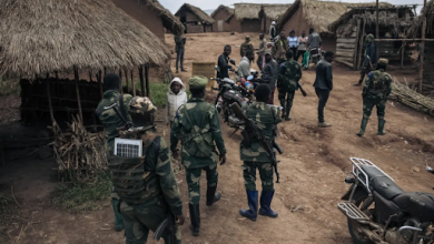 Soldiers in uniform gather in a village with thatched roofs, interacting with locals. Motorbikes are parked nearby.
