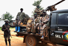 Soldiers in camouflage and gear with rifles sit on a truck in a rural setting, one is standing nearby. Trees and buildings in the background.