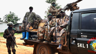 Soldiers in camouflage and gear with rifles sit on a truck in a rural setting, one is standing nearby. Trees and buildings in the background.
