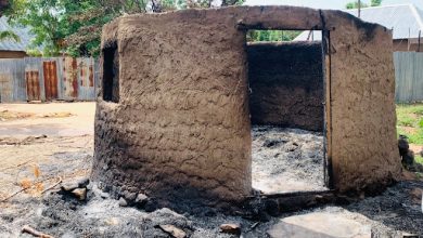 Round mud hut with an open doorway, surrounded by charred debris and sparse vegetation, against a backdrop of trees and metal fencing.