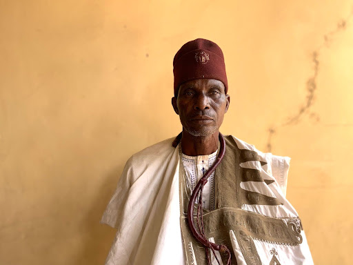 Man in traditional attire with a maroon cap stands against a plain, yellowish wall.