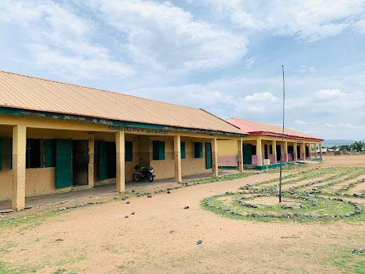A long building with a corrugated metal roof, open doors, and windows, set in a sandy area with sparse grass and small rocks.