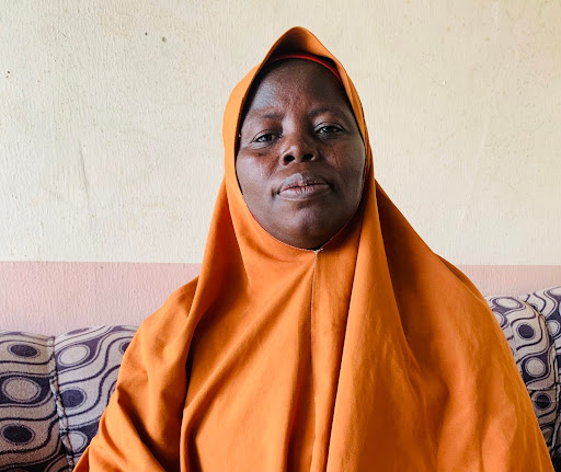 Person in an orange hijab sits on a patterned sofa, facing forward with a neutral expression.