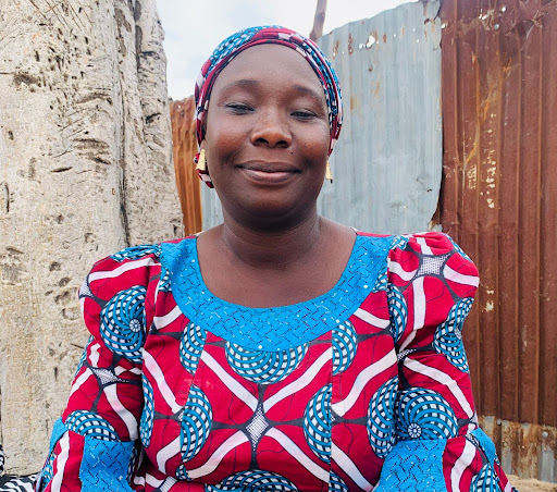 Smiling woman in a colorful patterned dress and headscarf, sitting outdoors with a rustic background.