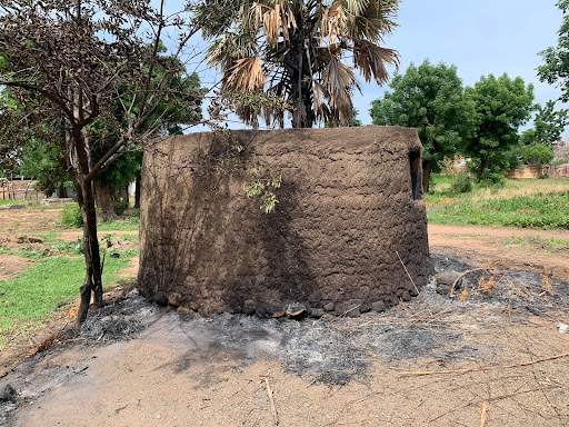 Round mud hut with a thatched roof, surrounded by charred ground and sparse trees in a rural setting.
