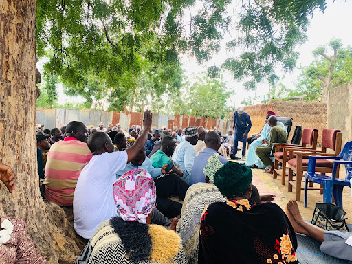 Community meeting outdoors under a tree, attendees seated and attentive, one person raising a hand.