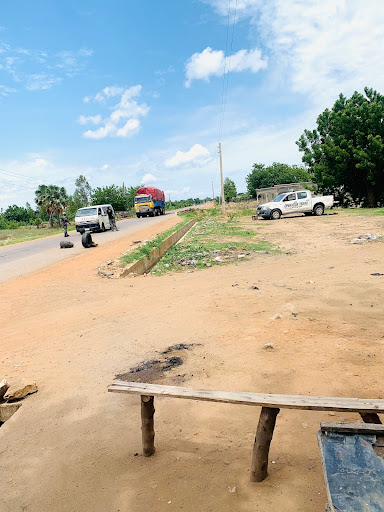 Rural road with parked vehicles, including a truck and a car. Dirt foreground, trees lining the road, and a partly cloudy sky above.