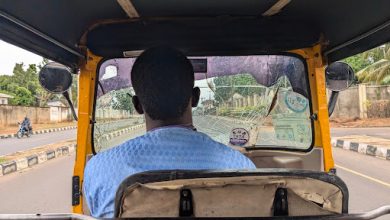 View from the back of a tuk-tuk, focusing on the driver, with a street scene visible through a cracked windshield.