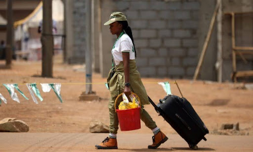A person in uniform walks while carrying a red bucket and pulling a suitcase along a street with buildings in the background.