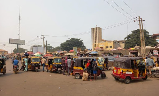 Busy street scene with multiple parked rickshaws, vendors, and people walking around under a clear sky. Buildings and power lines in the background.