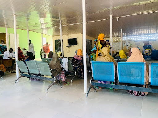 People seated in a waiting area with blue chairs and a TV on the wall, some standing, in a room with green accents and a wooden ceiling.