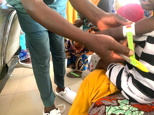 Child's arm being measured for growth with a tape in a clinic, surrounded by people.