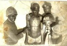 A group of men in uniform, some wearing helmets, surround a shirtless man in an old, worn photograph.
