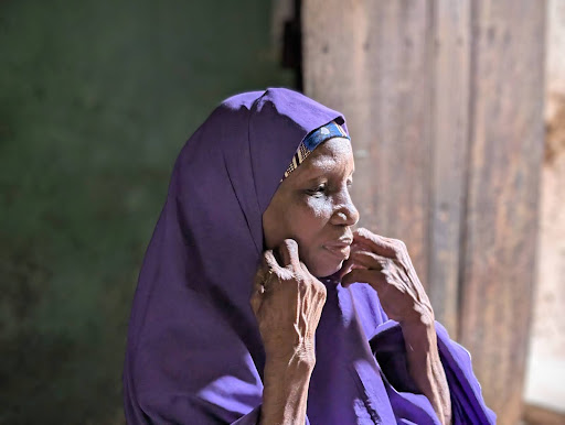 An elderly woman in a purple headscarf looks contemplatively out a window, with soft light illuminating her face and hands.