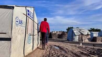 A person walks in a refugee camp with white tents and a muddy path under a blue sky.