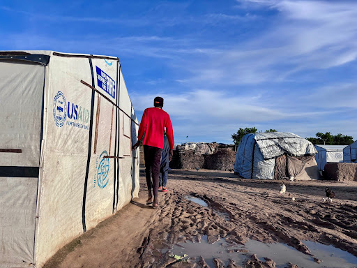 A person walks in a refugee camp with white tents and a muddy path under a blue sky.