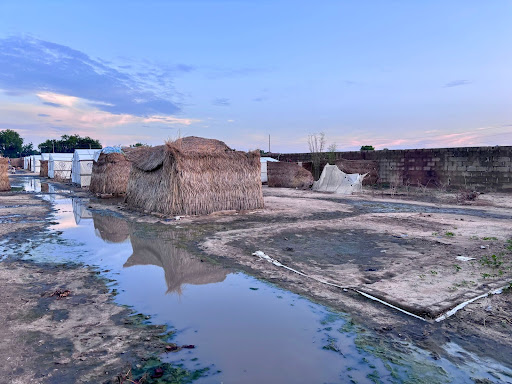 Straw huts in a rural area with puddles reflecting the sky, bordered by a concrete wall under a blue and cloudy evening sky.