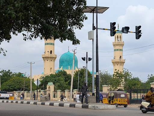 Street scene with traffic and motorcycles in front of a large mosque with a turquoise dome and two minarets.