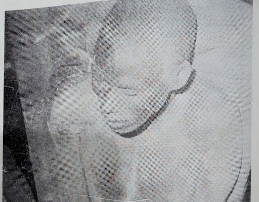 A grainy black-and-white photo of a person with a shaved head looking down, appearing seated or crouched.