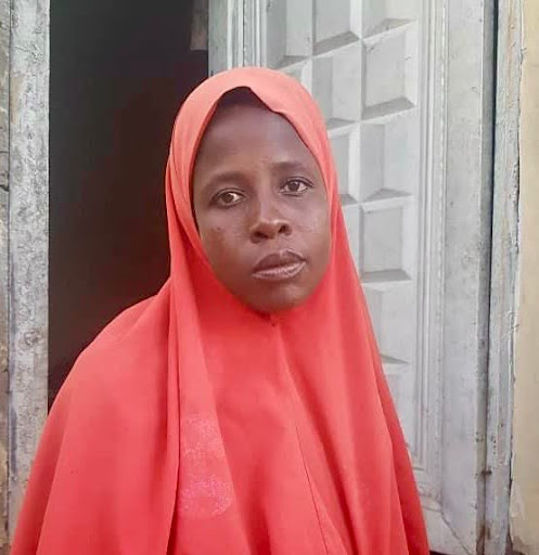 A woman in an orange headscarf stands in front of a decorative door, looking directly at the camera.
