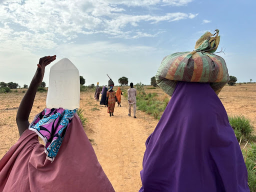 People in colorful clothing walk on a dirt path under a blue sky, with one carrying a water container.