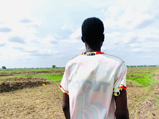 Person standing in a field, wearing a light-colored shirt with red and black accents, looking towards the horizon under a cloudy sky.