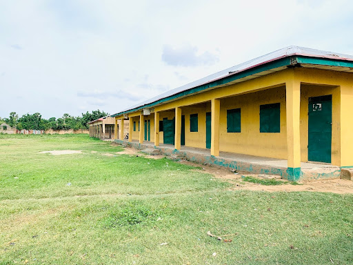 Long, yellow school building with green doors and windows, set on a grassy field under a partly cloudy sky.