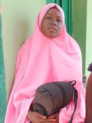 A person in a pink hijab holds a black bag, standing against a green wall.