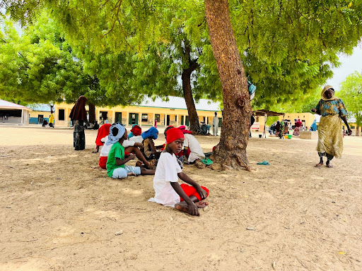 Children sitting under trees for shade on a sunny day, with a woman walking nearby and a building in the background.