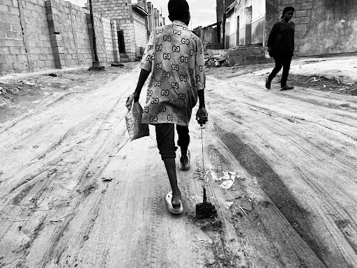 Child in a patterned shirt walks on a dirt road, pulling a toy. Another person walks in the opposite direction. Black and white photo.