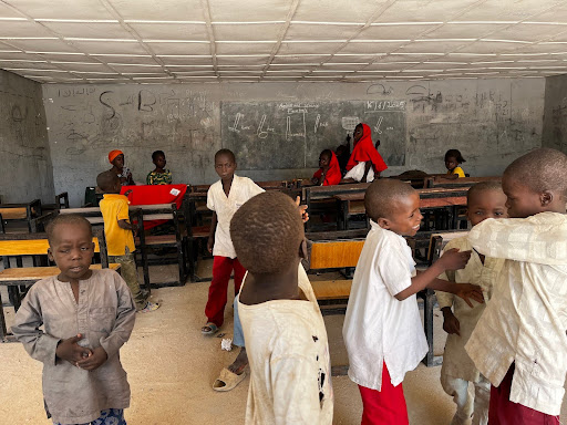 Children in a classroom, some gathered in groups talking and others sitting at desks.