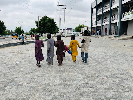 Five children walk hand in hand on a paved street near buildings, holding a blue balloon, cloudy sky above.
