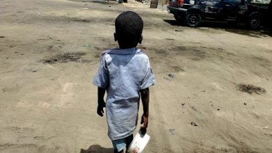 A child in a blue shirt walks on a sandy street, holding a white container. A car is parked in the background.