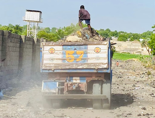 A person stands on top of a garbage-filled truck marked "57," on a dirt road with trees and a building in the background.