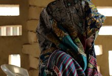 Person draped in vibrantly patterned fabric sits on a white chair, with light filtering through a textured brick wall behind them.