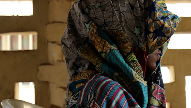 Person draped in vibrantly patterned fabric sits on a white chair, with light filtering through a textured brick wall behind them.