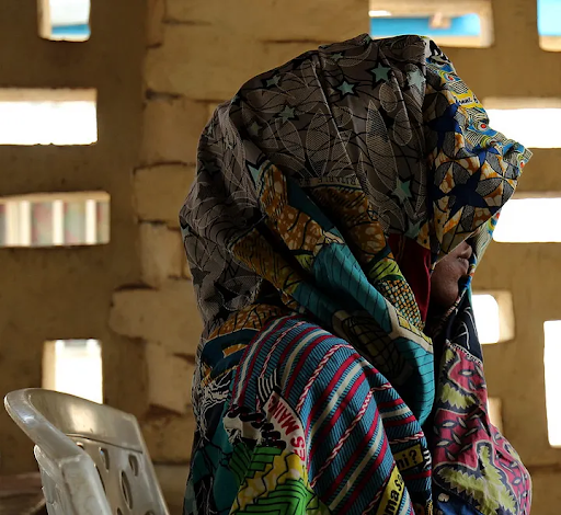 Person draped in vibrantly patterned fabric sits on a white chair, with light filtering through a textured brick wall behind them.