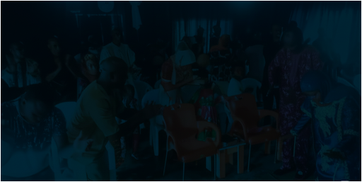 A group of people sitting and standing in a dimly lit room, some appear to be clapping. Brightly colored chairs are in the center.