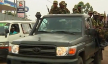 Military personnel ride in a green pickup truck on a busy street, surrounded by traffic, including a white van in the background.